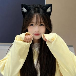 Puede incluir: Un par de orejas de gato negras con detalles interiores blancos sujetas a una diadema, combinadas con una sudadera de manga larga de color amarillo pálido con puños acanalados. Las orejas están hechas de un material suave y afelpado.
