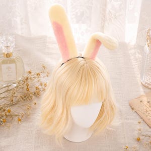 May include: A headband featuring two tall, plush rabbit ears in cream and soft pink, worn on a mannequin head with a shoulder-length, wavy blonde wig. The ears are upright, with one standing straight and the other slightly bent at the top.