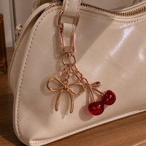 May include: A cream-colored handbag with a glossy finish, featuring a gold-toned metal charm attached to the handle. The charm consists of a delicate gold bow and two shiny, translucent red cherry beads hanging from a short gold chain link.