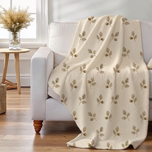 May include: A cream-colored throw blanket featuring a repeating pattern of small brown acorns and green oak leaves. The blanket is draped over a sofa, showcasing the detailed botanical print across the fabric surface with a soft, textured appearance.
