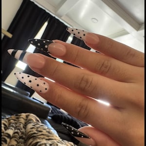 Op de afbeelding: Lange, stiletto-vormige kunstnagels in een French manicure-stijl met een nude basis. De uiteinden zijn versierd met zwarte stippen op een witte of zwarte achtergrond, en één nagel heeft zwarte horizontale strepen op een witte punt.
