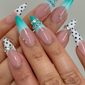 Könnte beinhalten: Lange, stilettoförmige künstliche Fingernägel mit verschiedenen Designs. Einige Nägel haben eine weiße Basis mit schwarzen Punkten, während andere eine türkisfarbene bis weiße Farbverlauf-Spitze aufweisen. Mehrere Nägel sind mit Strasssteinen verziert.