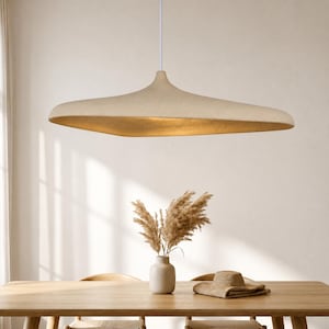 May include: A wide, saucer-shaped pendant light with a textured, beige exterior and a warm, glowing interior. The fixture is suspended from a thin white cord, featuring an organic, asymmetrical silhouette that casts a soft light downwards.