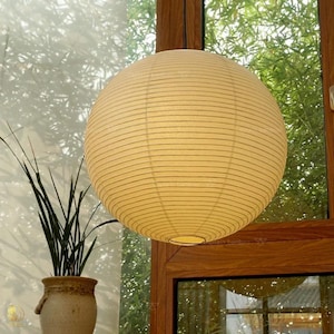 May include: A spherical paper lantern with a ribbed texture and a warm off-white color. The light fixture features a circular opening at the bottom and is suspended in front of a window with a wooden frame and green foliage visible outside.