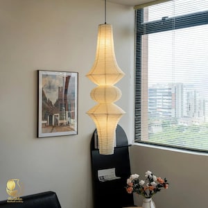 May include: A tall, off-white paper pendant light fixture with a ribbed texture and a stacked, multi-tiered silhouette. The lamp features a narrow top section, a central rounded bulbous segment, and a wider base, suspended by a thin black cord.