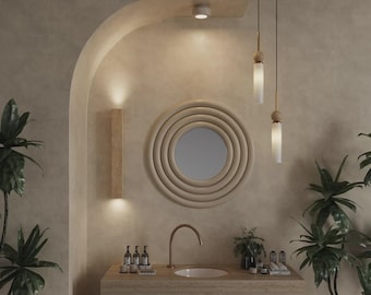 Könnte beinhalten: Ein Badezimmer-Waschtischbereich mit einem runden Spiegel mit konzentrischen Ringrahmen, einem geschwungenen Kupferwasserhahn und einem rechteckigen Waschbeckenunterschrank aus Holz. Zwei Pendelleuchten mit zylindrischen Glasschirmen und zwei Wandleuchten sorgen für Beleuchtung.