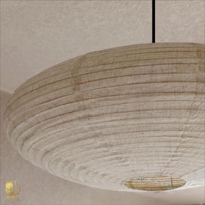 Pode incluir: Uma luminária pendente grande e redonda de papel em tom off-white com textura canelada. A cúpula apresenta uma estrutura interna de arame visível e está suspensa por um fio preto fino preso ao teto, criando um visual suave e difuso.