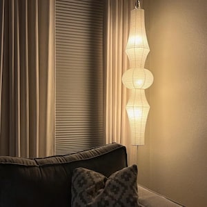 Op de afbeelding: Een hoge, verticale vloerlamp met drie gestapelde, gebroken witte papieren lantaarnkappen met een geribbelde textuur. De lamp verspreidt een warme, zachte gloed van binnenuit, die de cilindrische en afgeronde delen van de papieren structuur verlicht.