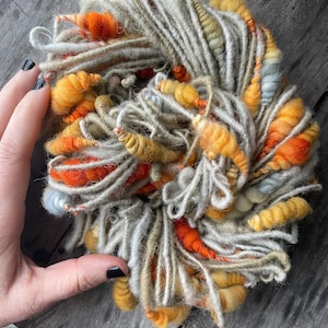 Könnte beinhalten: Ein Knäuel handgesponnener Wolle in Orange-, Gelb- und Grautönen. Die Wolle hat eine dicke, strukturierte Optik, mit Bereichen, die dichter oder lockerer gewickelt sind. Ideal zum Stricken, Weben oder für andere Textilkunstprojekte, bietet diese Wolle eine einzigartige Textur.