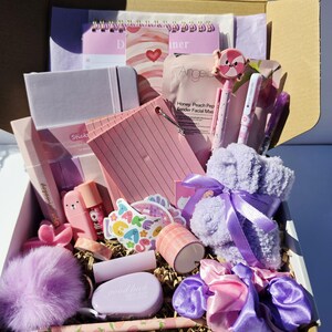 May include: A gift box overflowing with pink and purple items. Contents include a planner, notebook, pens, stickers, lip gloss, a pom-pom, scrunchies, and fluffy socks tied with ribbon. The box is lined with tissue paper and decorative paper.