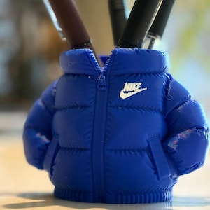 May include: A blue pen holder in the shape of a puffer jacket, featuring a zip and the Nike logo in white. Several pens are inside the holder. The pen holder is on a light-coloured surface.