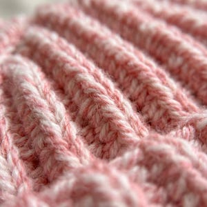 May include: A close-up view of a knitted fabric featuring a ribbed texture. The yarn is a blend of soft pink and white fibers, creating a variegated appearance throughout the knit pattern. The stitches are clearly visible, showing a consistent vertical.