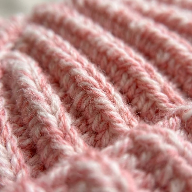 May include: A close-up view of a knitted fabric featuring a ribbed texture. The yarn is a blend of soft pink and white fibers, creating a variegated appearance throughout the knit pattern. The stitches are clearly visible, showing a consistent vertical.