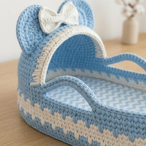 May include: A light blue and white crocheted bassinet with a hood featuring two rounded ears and a central white bow. The structure has a woven texture with a zigzag pattern along the base and a light blue and white chevron fabric lining inside.