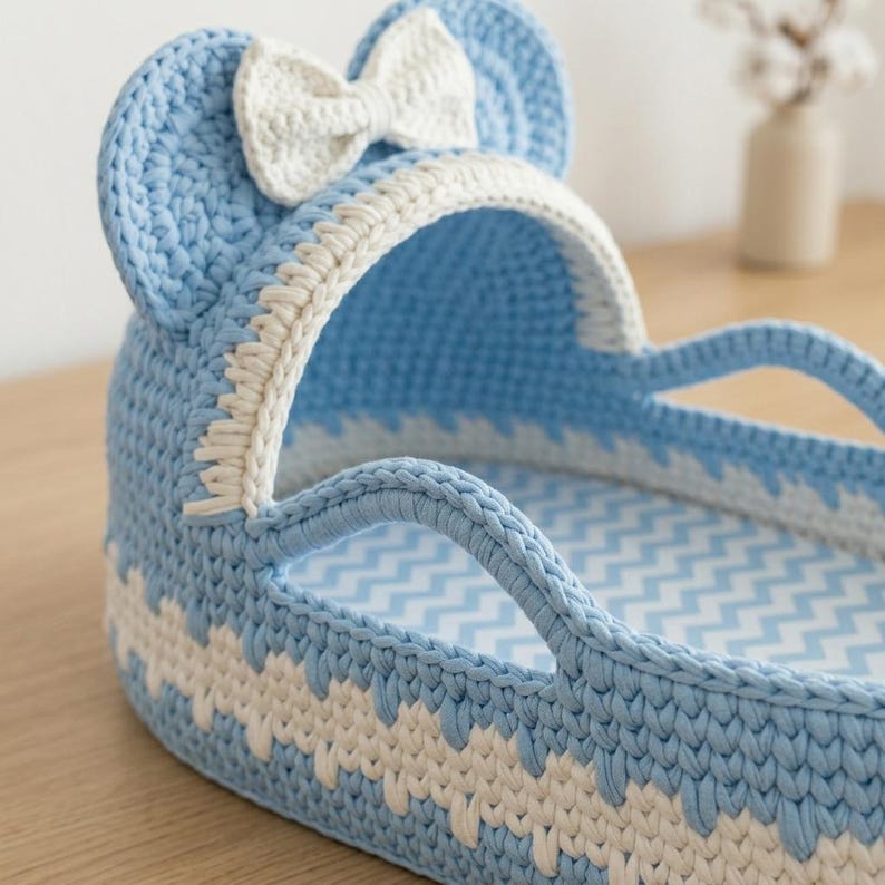 May include: A light blue and white crocheted bassinet with a hood featuring two rounded ears and a central white bow. The structure has a woven texture with a zigzag pattern along the base and a light blue and white chevron fabric lining inside.