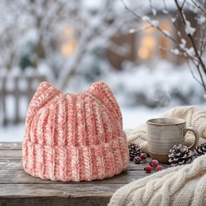 May include: A chunky knit beanie in a variegated peach and white yarn, featuring two pointed cat-ear shapes at the top and a wide ribbed cuff. The hat is displayed on a wooden surface alongside a ceramic mug and a cream-colored knit fabric.
