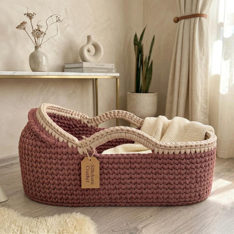 Peut inclure: Un couffin en crochet mauve profond avec un bord et des anses de couleur cr&egrave;me. Une couverture en tricot couleur cr&egrave;me est plac&eacute;e &agrave; l'int&eacute;rieur. Une &eacute;tiquette en papier beige attach&eacute;e &agrave; l'anse porte le texte imprim&eacute; Stitchora Crochet.
