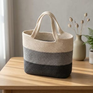 May include: A crochet tote bag featuring a horizontal colour-blocked design with three distinct sections. The top section is cream, the middle section is light grey, and the bottom section is dark charcoal grey. The bag has two integrated handles.