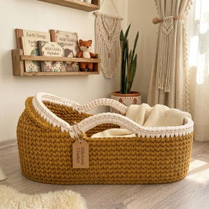 Peut inclure: Un couffin pour b&eacute;b&eacute; en crochet jaune moutarde avec un bord tress&eacute; et des anses de couleur cr&egrave;me. Une &eacute;tiquette rectangulaire en carton attach&eacute;e &agrave; l'anse porte le texte Stitchora Crochet. Une couverture en tricot cr&egrave;me est pos&eacute;e &agrave; l'int&eacute;rieur.