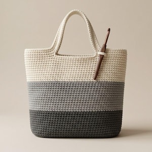 May include: A crochet tote bag featuring a color-blocked design with horizontal stripes in cream, light gray, and dark charcoal gray. The bag has two integrated handles and a brown crochet hook tucked into the top edge of the cream section.