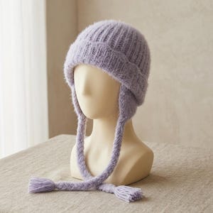 Puede incluir: Un gorro de punto difuso en color violeta claro con textura acanalada y orejeras. El gorro cuenta con dos largas tiras laterales trenzadas que cuelgan, cada una terminada con una pequeña borla en el extremo. El material tiene un aspecto suave.