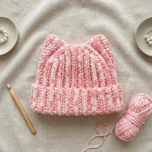 May include: A pink and white variegated yarn beanie with cat-ear points, featuring a ribbed knit texture and a folded brim. The hat is accompanied by a wooden crochet hook and a matching ball of yarn, with two small white pearl-like beads nearby.