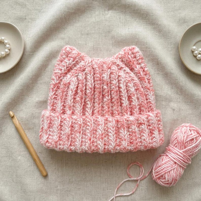 May include: A pink and white variegated yarn beanie with cat-ear points, featuring a ribbed knit texture and a folded brim. The hat is accompanied by a wooden crochet hook and a matching ball of yarn, with two small white pearl-like beads nearby.