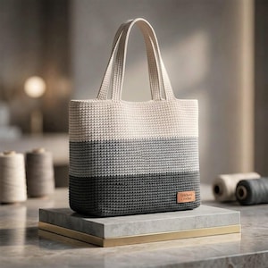 May include: A crochet tote bag featuring a three-tone color block design in cream, light gray, and dark charcoal gray. The bag has two integrated handles and a small rectangular brown leather-like tag on the bottom right corner with the text Stitchora Crochet.