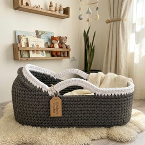 Peut inclure: Un couffin pour b&eacute;b&eacute; ovale en crochet gris fonc&eacute; avec un bord &eacute;pais en crochet blanc et des poign&eacute;es int&eacute;gr&eacute;es. Une couverture en tricot cr&egrave;me est pos&eacute;e &agrave; l'int&eacute;rieur. Une &eacute;tiquette en papier brun porte le texte Stitchora Crochet.
