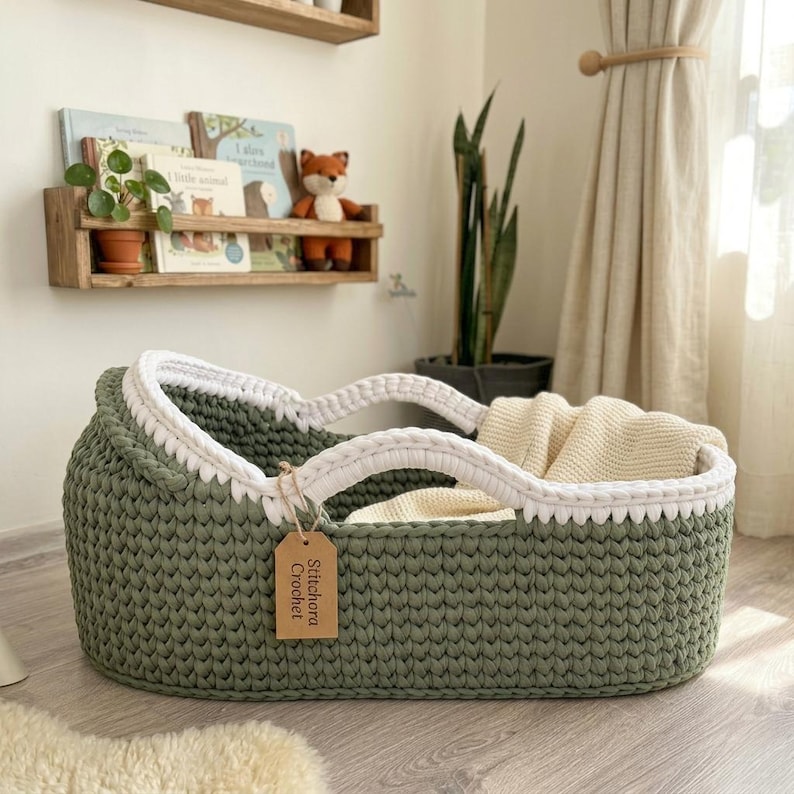 Peut inclure: Un couffin pour b&eacute;b&eacute; en crochet de forme ovale vert sauge, dot&eacute; d'une bordure tress&eacute;e blanche &eacute;paisse sur le dessus et de deux anses int&eacute;gr&eacute;es. Une couverture en tricot cr&egrave;me est plac&eacute;e &agrave; l'int&eacute;rieur et une &eacute;tiquette en papier indique Stitchora Crochet.