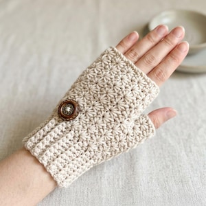 Puede incluir: Un guante sin dedos de color beige hecho de hilo de ganchillo con un patrón de puntos en forma de estrella. El guante incluye una sección de puño acanalado y un botón circular marrón oscuro con un acento blanco central en el lateral.