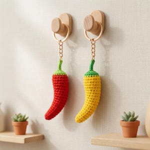 May include: Two crochet chilli pepper keychains hanging from light wood wall hooks. One pepper is bright red and the other is vibrant yellow. Each keychain features a metal ring and chain attached to the top of the textured, handmade pepper shape.