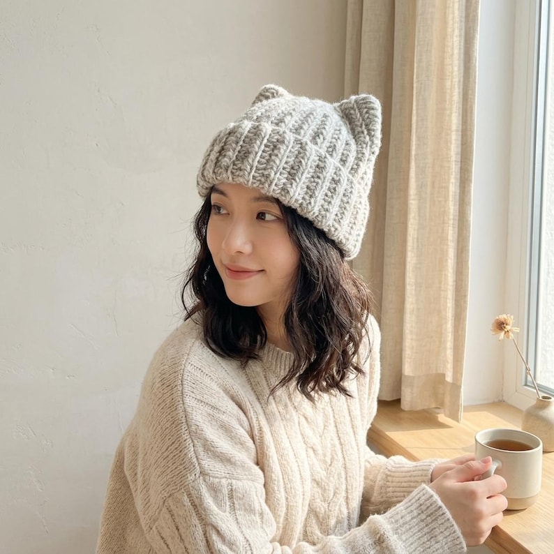 May include: A light gray knit beanie featuring two small pointed cat-ear shapes at the top. The hat is constructed with a thick ribbed texture and a folded brim, crafted from a soft yarn material that provides a cozy and structured appearance.