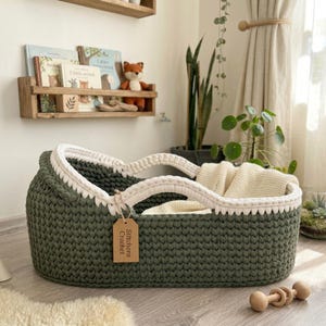 Peut inclure: Un couffin pour b&eacute;b&eacute; en crochet vert olive fonc&eacute; de forme ovale, dot&eacute; d'un bord tress&eacute; &eacute;pais de couleur cr&egrave;me et de poign&eacute;es assorties. Une &eacute;tiquette en papier brun avec le texte Stitchora Crochet est fix&eacute;e sur le c&ocirc;t&eacute;. Une couverture cr&egrave;me est &agrave; l'int&eacute;rieur.