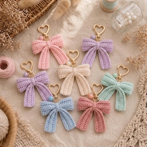 May include: A collection of seven crochet bow keychains in pastel colours including pink, lavender, cream, light blue, and mint green. Each bow features a gold-toned heart-shaped clasp and a small round bead attached to the top of the bow.