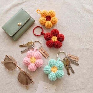 May include: A sage green leather wallet, brown sunglasses, and four crocheted flower keychains in yellow, red, pink, and light blue. Each keychain features a metal ring and a small wooden tag engraved with the text MADE WITH LOVE.