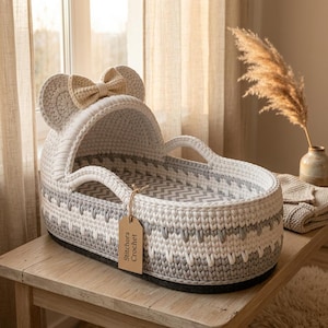 May include: A white and gray crocheted bassinet with a hood featuring two rounded ears and a cream-colored bow. The interior has a gray and white chevron pattern. A brown paper tag attached to the handle reads Stitchora Crochet in black text.