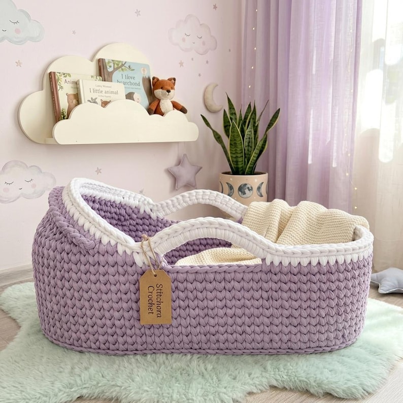 Peut inclure: Un couffin pour b&eacute;b&eacute; en crochet lavande avec un bord blanc et deux anses int&eacute;gr&eacute;es. Une &eacute;tiquette rectangulaire beige portant le texte Stitchora Crochet est attach&eacute;e sur le c&ocirc;t&eacute;. &Agrave; l'int&eacute;rieur, une couverture en tricot cr&egrave;me est pli&eacute;e.