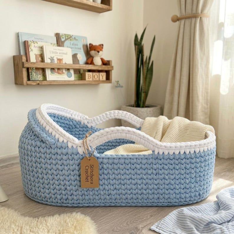 Peut inclure: Un panier ovale en crochet bleu clair avec un bord tress&eacute; blanc &eacute;pais et deux anses int&eacute;gr&eacute;es. Une &eacute;tiquette rectangulaire en papier brun est attach&eacute;e sur le c&ocirc;t&eacute; avec le texte Stitchora Crochet imprim&eacute; en noir. Une couverture cr&egrave;me est &agrave; l'int&eacute;rieur.