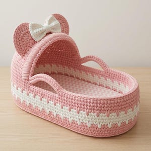 May include: A pink and white crocheted bassinet featuring a hood with two rounded ears and a central white bow. The oval-shaped basket has a textured stitch pattern with a white zigzag band around the middle and two integrated side handles.