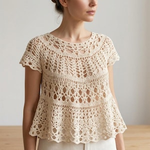 May include: A cream-coloured crochet top featuring an intricate openwork pattern with a round neckline and short sleeves. The garment has a flared, peplum-style silhouette with detailed lace-like stitching throughout the bodice and hemline.