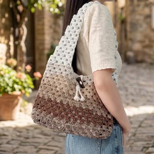 Puede incluir: Un bolso de hombro de ganchillo con patrón de granny square y un degradado de color que va del azul claro en la parte superior al marrón oscuro en la base. El bolso incluye un cierre de cordón blanco con un botón oscuro y una correa ancha.