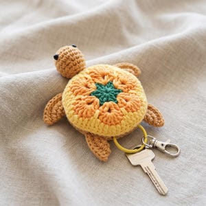 Peut inclure: Un porte-clés tortue en crochet avec une tête et des membres couleur beige, et une carapace composée de fils jaune, orange et vert foncé formant un motif floral. Un anneau jaune relie la tortue à un mousqueton en métal argenté et une clé.