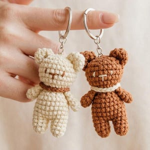 Può includere: Due piccoli portachiavi a forma di orsetto all'uncinetto, uno color crema e uno marrone. Ogni orsetto presenta un viso ricamato semplice, orecchie arrotondate e un colletto in filato di colore contrastante. Le figure sono fissate ad anelli metallici.
