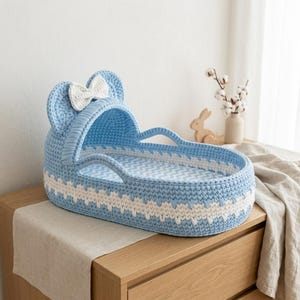 May include: A light blue crocheted baby bassinet featuring a white crocheted bow on the hood, a white zigzag pattern along the base, and a light blue and white chevron patterned mattress pad. The structure includes two integrated side handles.