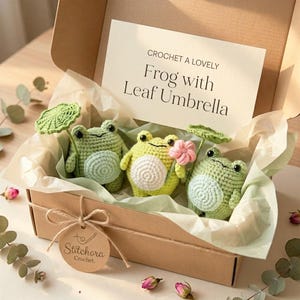 May include: A cardboard gift box containing three crocheted frog figures with light green bodies and white bellies. Two frogs hold green crocheted leaf umbrellas, and one holds a pink flower. A card inside reads: CROCHET A LOVELY Frog with Leaf Umbrella.