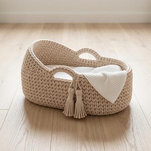 May include: A beige oval-shaped crochet basket with integrated handles and two decorative hanging tassels on the side. The basket features a thick, textured knit pattern and contains a white waffle-weave fabric lining inside the base.