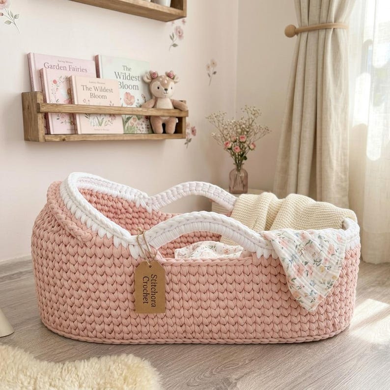 Peut inclure: Un couffin pour b&eacute;b&eacute; ovale en crochet rose clair avec une bordure tress&eacute;e blanche &eacute;paisse et deux anses solides. Une &eacute;tiquette en carton beige marqu&eacute;e Stitchora Crochet est attach&eacute;e sur le c&ocirc;t&eacute;. Une couverture cr&egrave;me et un tissu floral sont &agrave; l'int&eacute;rieur.