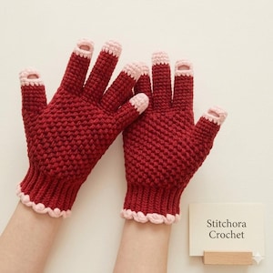 Könnte beinhalten: Ein Paar dunkelrote Häkelhandschuhe mit hellrosa Besatz an den Bündchen und Fingerspitzen. Die Handschuhe haben ein strukturiertes Maschenmuster und offene Spitzen an Daumen, Zeige- und Mittelfinger. Eine Karte trägt die Aufschrift Stitchora Crochet.