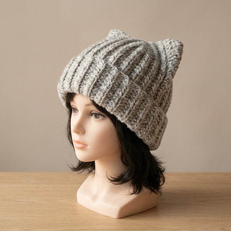 May include: A light gray, chunky knit beanie with a ribbed texture and two pointed cat-ear shapes at the top. The hat is crafted from a thick, heathered yarn and features a wide, folded brim that sits snugly around the head of the mannequin.